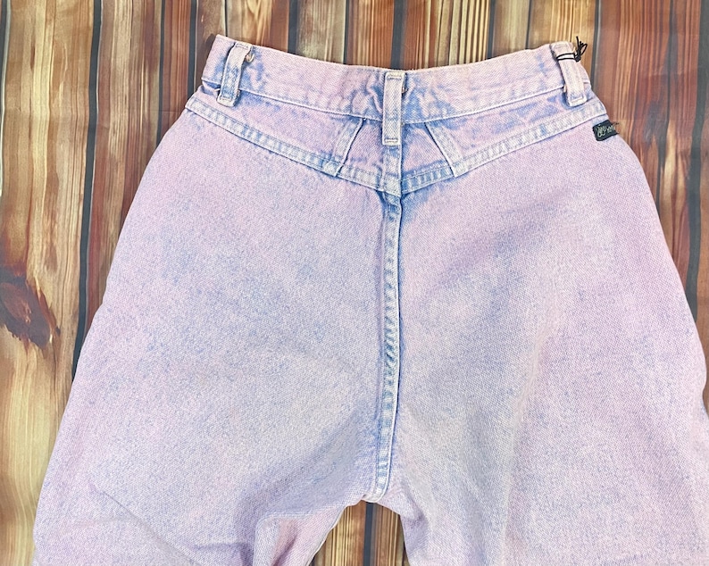 Pu&ograve; includere: Jeans vintage a vita alta in un lavaggio acido lilla chiaro. I jeans presentano un classico design a cinque tasche, passanti per cintura e un carr&egrave; a forma di V sul retro. Il denim ha un aspetto sbiadito e usurato, con una piccola etichetta nera sul retro.