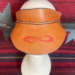 May include: A tan leather visor with the word "INFINITY" in red. The visor has a curved shape and is stitched with black thread. The visor is on a white mannequin head.