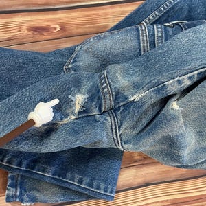 May include: Close-up of blue denim jeans with distressed details, including frayed edges and small holes. The jeans are folded, showcasing the texture and wear of the fabric. The stitching is visible, highlighting the construction of the garment.