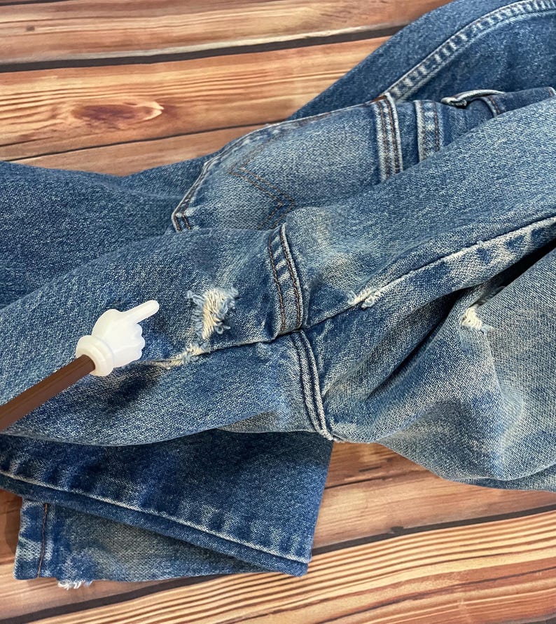 May include: Close-up of blue denim jeans with distressed details, including frayed edges and small holes. The jeans are folded, showcasing the texture and wear of the fabric. The stitching is visible, highlighting the construction of the garment.