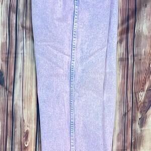 Pu&ograve; includere: Jeans in denim lavato all'acido di colore viola chiaro. I jeans hanno un classico design a cinque tasche con chiusura con bottone e cerniera. Il denim ha un aspetto sbiadito e vintage. I jeans sono esposti su una superficie di legno.