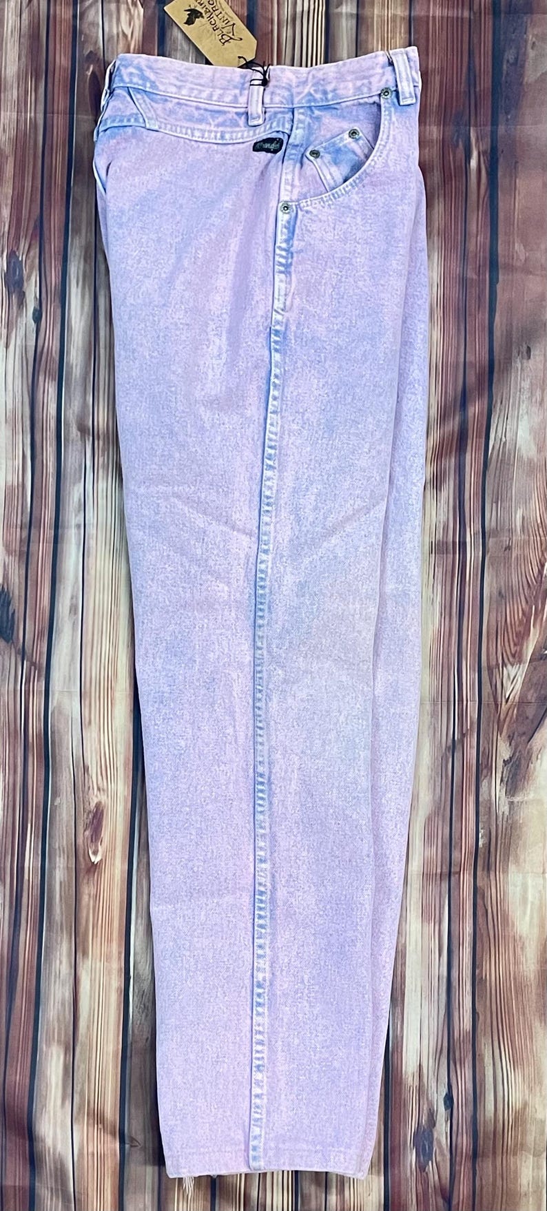 Pu&ograve; includere: Jeans in denim lavato all'acido di colore viola chiaro. I jeans hanno un classico design a cinque tasche con chiusura con bottone e cerniera. Il denim ha un aspetto sbiadito e vintage. I jeans sono esposti su una superficie di legno.