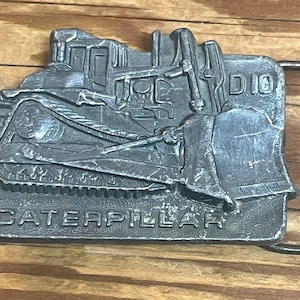 Könnte beinhalten: Silberfarbene Gürtelschnalle mit einem detaillierten Relief eines Caterpillar-Bulldozers. Das Wort "CATERPILLAR" ist unten eingeprägt. Die Schnalle hat eine rechteckige Form mit einer Metallschlaufe.