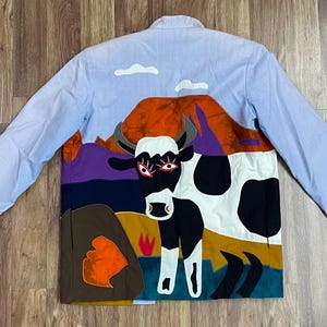 May include: Light blue button-up jacket with a colorful patchwork design. The back features a cow with red eyes, a mountain scene, and a brown rock formation. The jacket has long sleeves and a classic collar.