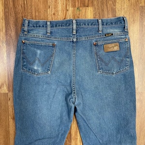Może przedstawiać: Dżinsy z niebieskiego denimu z brązową sk&oacute;rzaną metką Wrangler na tylnej kieszeni.