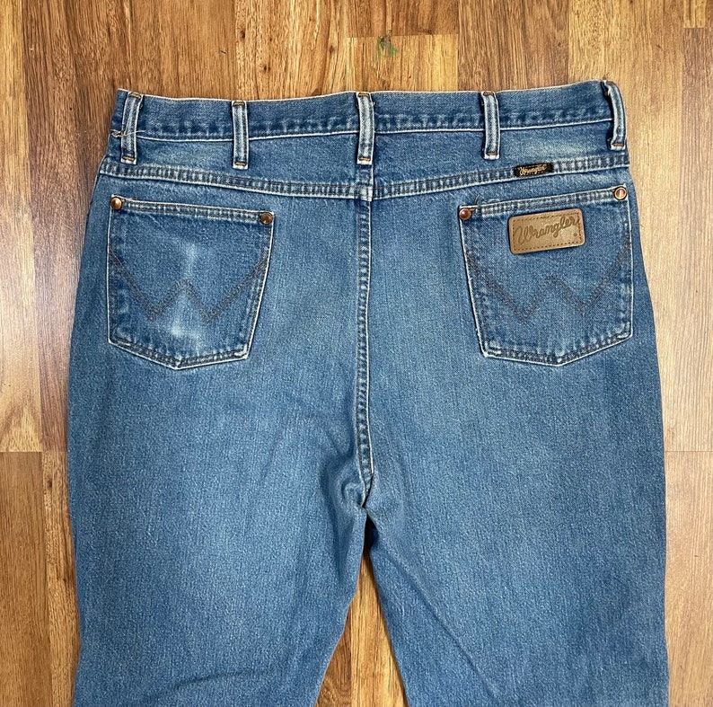 Może przedstawiać: Dżinsy z niebieskiego denimu z brązową sk&oacute;rzaną metką Wrangler na tylnej kieszeni.