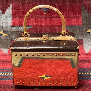 Könnte beinhalten: Eine rechteckige Vintage-Handtasche mit braunem Oberteil und goldfarbenen Metallseiten. Die Tasche hat einen goldenen Griff und ist mit kleinen, goldfarbenen Nieten verziert. Die Tasche steht auf einer rot-braun gemusterten Oberfläche.