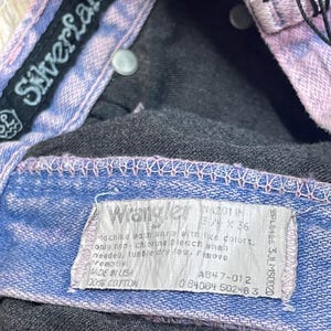 Pu&ograve; includere: Primo piano di un paio di jeans in denim con finiture blu chiaro e viola. I jeans hanno un'etichetta "Wrangler" e un'etichetta del marchio "SilverLake". L'etichetta indica che i jeans sono 5 3/4 x 36 pollici (circa 14.6 x 91.4 cm).