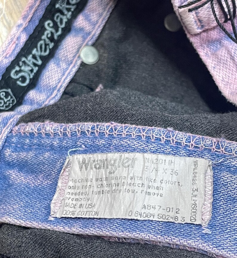 Pu&ograve; includere: Primo piano di un paio di jeans in denim con finiture blu chiaro e viola. I jeans hanno un'etichetta "Wrangler" e un'etichetta del marchio "SilverLake". L'etichetta indica che i jeans sono 5 3/4 x 36 pollici (circa 14.6 x 91.4 cm).