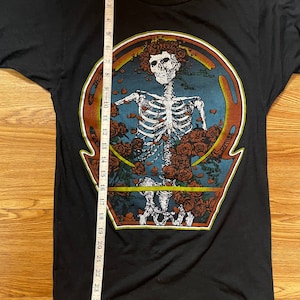 Puede incluir: Camiseta negra con un estampado gr&aacute;fico de un esqueleto con rosas alrededor de la cabeza. El esqueleto est&aacute; dentro de un c&iacute;rculo con fondo azul y borde naranja. El texto "Steal Your Face" est&aacute; impreso en la parte inferior del gr&aacute;fico.