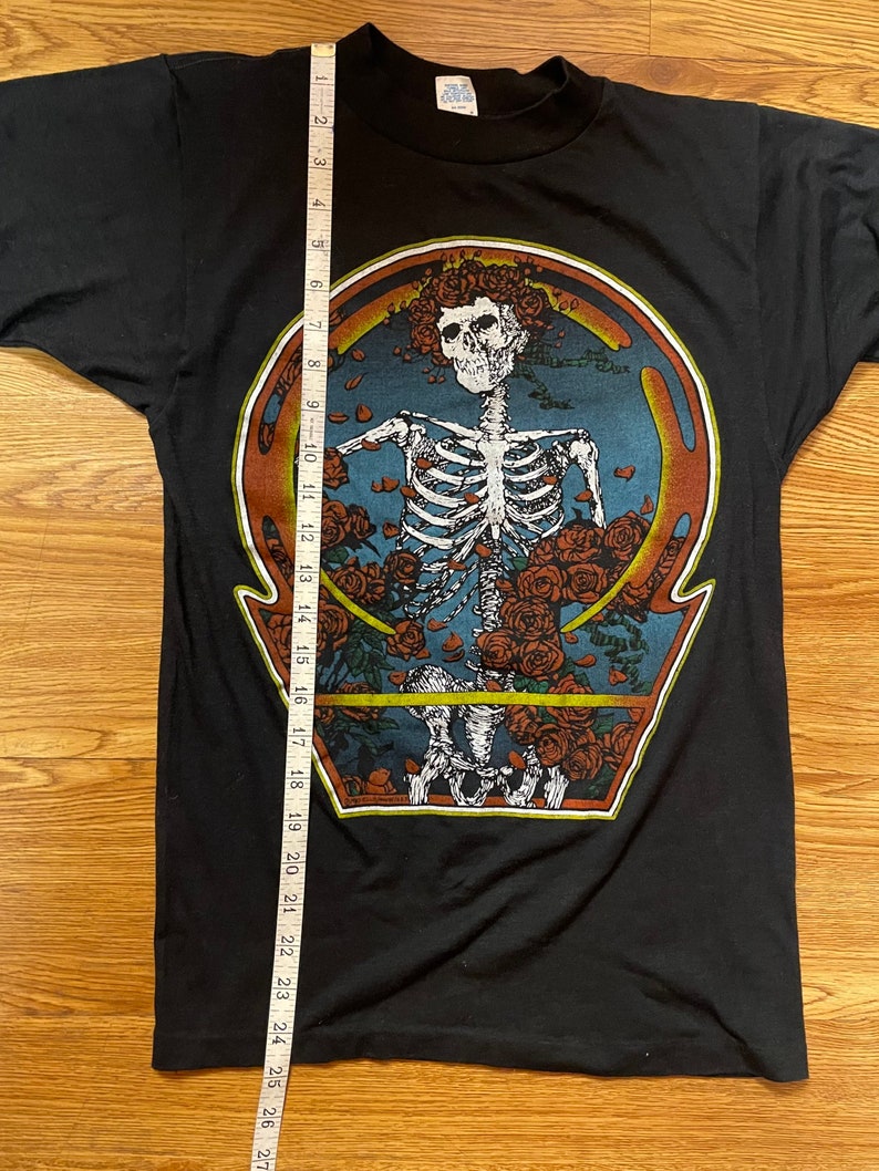 Puede incluir: Camiseta negra con un estampado gr&aacute;fico de un esqueleto con rosas alrededor de la cabeza. El esqueleto est&aacute; dentro de un c&iacute;rculo con fondo azul y borde naranja. El texto "Steal Your Face" est&aacute; impreso en la parte inferior del gr&aacute;fico.