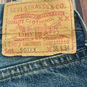 Puede incluir: Primer plano de una etiqueta de vaqueros. La etiqueta es de color beige con letras y gráficos rojos, incluido el texto "LEVI STRAUSS & CO." y "ORIGINAL RIVETED". La etiqueta también indica la talla: W38 L34. Los vaqueros son azules.