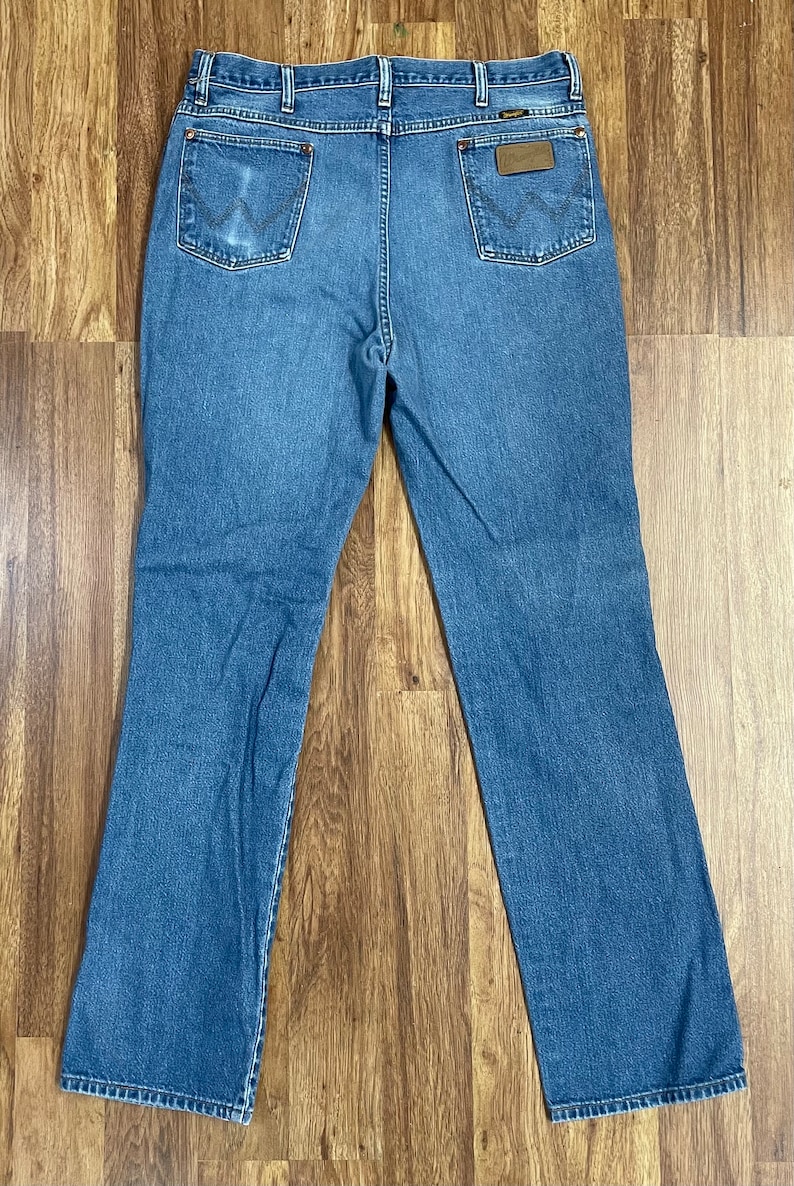 Może przedstawiać: Dżinsy z niebieskiego denimu z brązową sk&oacute;rzaną naszywką na tylnej kieszeni z napisem "Wrangler".