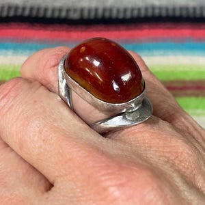 Puede incluir: Un anillo de plata con una gran piedra de ámbar ovalada y lisa de tono marrón rojizo intenso. La banda de metal tiene un acabado ancho y pulido que se curva alrededor de la base del engaste, proporcionando un soporte seguro para la gema brillante.