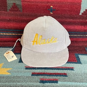 May include: A light gray corduroy baseball cap with the word "Alaska" embroidered in yellow script. The cap has a curved brim and a small tag attached to the side.