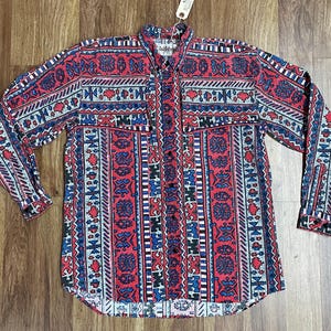 Op de afbeelding: Een lang mouw overhemd met knopen en een kleurrijk geometrisch patroon in tinten rood, blauw en wit. Het overhemd heeft een puntige kraag en een knoopsluiting aan de voorkant.