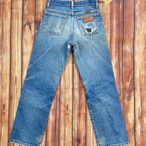 May include: Vintage blue denim Wrangler jeans with a distressed look. Features a classic five-pocket design, a Wrangler patch, and a small tear on the back pocket. The jeans are displayed on a wooden surface.