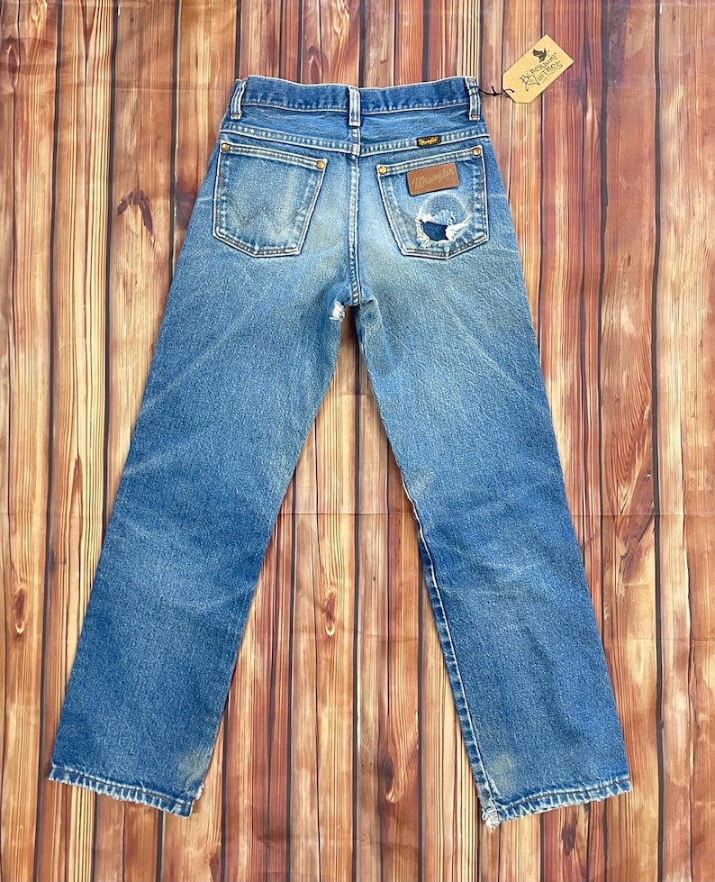 May include: Vintage blue denim Wrangler jeans with a distressed look. Features a classic five-pocket design, a Wrangler patch, and a small tear on the back pocket. The jeans are displayed on a wooden surface.