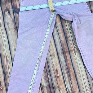 Pu&ograve; includere: Jeans di denim viola chiaro con un classico design a cinque tasche. I jeans sono misurati con un righello, mostrando una misura della vita di circa 38 cm e un'interno gamba di circa 76 cm. I jeans sono appoggiati su una superficie di legno.