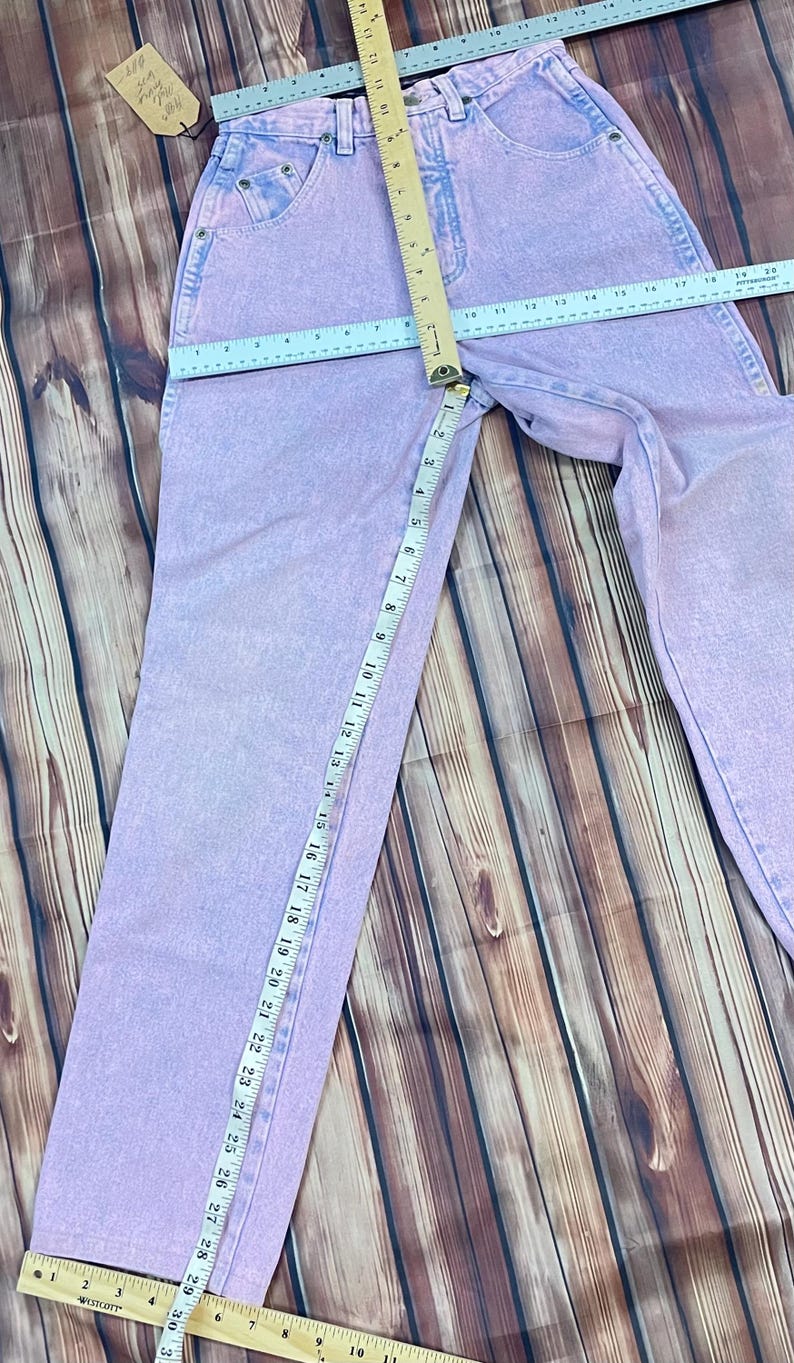 Pu&ograve; includere: Jeans di denim viola chiaro con un classico design a cinque tasche. I jeans sono misurati con un righello, mostrando una misura della vita di circa 38 cm e un'interno gamba di circa 76 cm. I jeans sono appoggiati su una superficie di legno.