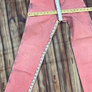 May include: Pink denim jeans with a straight leg, measured with a ruler. The waist measures approximately 15 inches across. The inseam is approximately 29 inches. The jeans are laid out on a wooden surface.