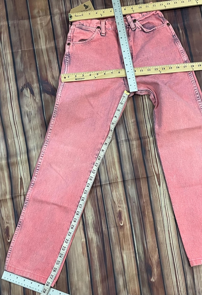 May include: Pink denim jeans with a straight leg, measured with a ruler. The waist measures approximately 15 inches across. The inseam is approximately 29 inches. The jeans are laid out on a wooden surface.