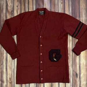 May include: A maroon cardigan sweater with a V-neck, button closure, and two front pockets. The left sleeve has three black stripes. A large black and gold letter "G" patch is on the left side. The tag reads "ALL N TIME".