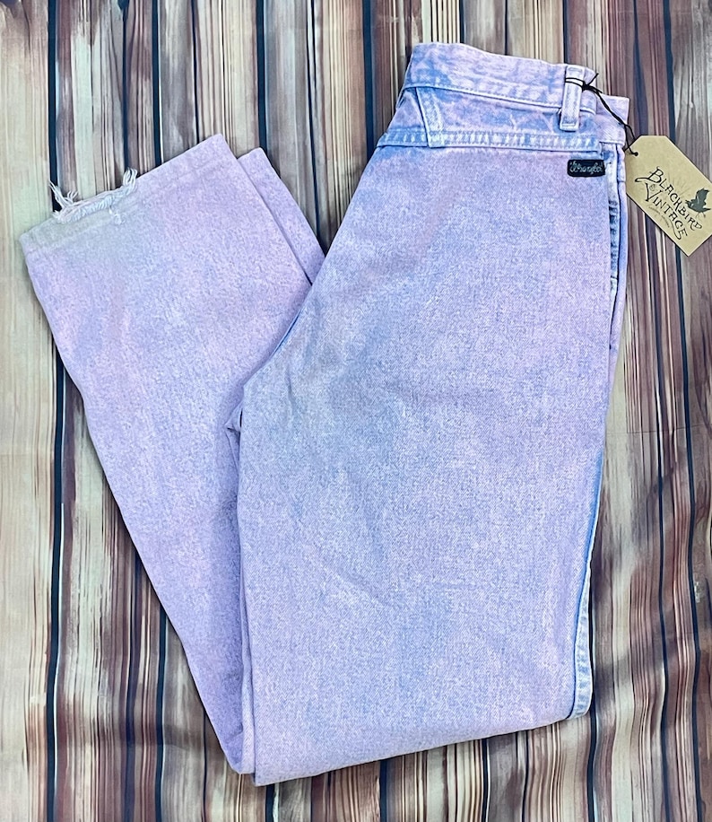 Pu&ograve; includere: Jeans Wrangler viola chiaro con effetto sbiadito. I jeans hanno un classico design a cinque tasche e una toppa con il logo Wrangler. &Egrave; attaccata un'etichetta Blackbird Vintage. I jeans sono appoggiati su una superficie di legno.