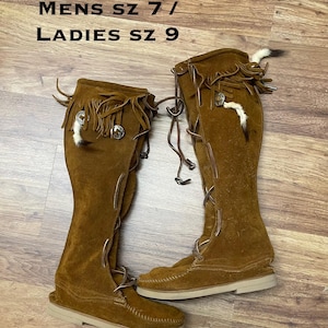Puede incluir: Un par de botas altas de ante marrón con flecos y botones plateados decorativos. Las botas tienen un diseño con cordones y una suela de color claro. El texto "MENS SZ 7 / LADIES SZ 9" es visible en la parte superior.
