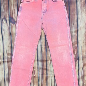 May include: Pink denim jeans with a classic five-pocket design. The jeans have a faded, vintage look with a straight leg cut. Features include copper rivets and a button closure. The jeans are displayed on a wooden surface.