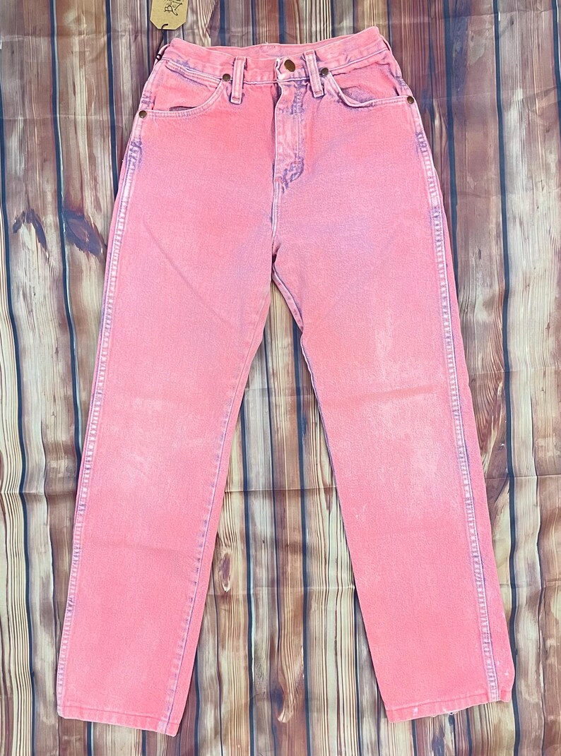 May include: Pink denim jeans with a classic five-pocket design. The jeans have a faded, vintage look with a straight leg cut. Features include copper rivets and a button closure. The jeans are displayed on a wooden surface.