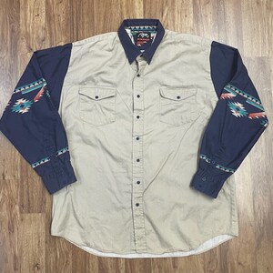 May include: A beige long-sleeve button-down shirt with navy blue sleeves featuring a geometric pattern. The shirt has two chest pockets and a label that reads "Roper Western Wear".