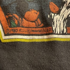 Puede incluir: Primer plano de una camiseta negra con un estampado gr&aacute;fico de un personaje de dibujos animados. El personaje lleva un sombrero verde y tiene la cara marr&oacute;n. El gr&aacute;fico est&aacute; rodeado por un borde blanco y amarillo.