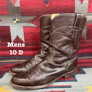 Op de afbeelding: Een paar donkerbruine leren cowboylaarzen met een halflange schacht, ronde neuzen en treklussen aan de zijkanten. De laarzen zijn voorzien van de tekst Mens 10 D, wat de maat en breedte aangeeft. Het leer heeft een gladde afwerking met stiksels.