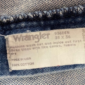 Może przedstawiać: Biała metka pielęgnacyjna dżins&oacute;w Wrangler z tekstem "936DEN 38 x 36", "Machine wash hot and inside out first three times with like colors, tumble dry", "Made in USA" i "100% Cotton".