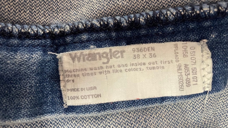 Może przedstawiać: Biała metka pielęgnacyjna dżins&oacute;w Wrangler z tekstem "936DEN 38 x 36", "Machine wash hot and inside out first three times with like colors, tumble dry", "Made in USA" i "100% Cotton".