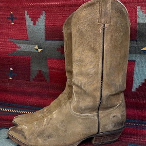 May include: A pair of worn, brown suede cowboy boots. The boots have decorative stitching, a pointed toe, and a stacked heel. A black line runs down the back of each boot. The boots are set against a patterned rug.