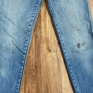 May include: A pair of blue denim jeans with visible wear and tear. The jeans have a faded appearance and are laid flat on a wooden surface.