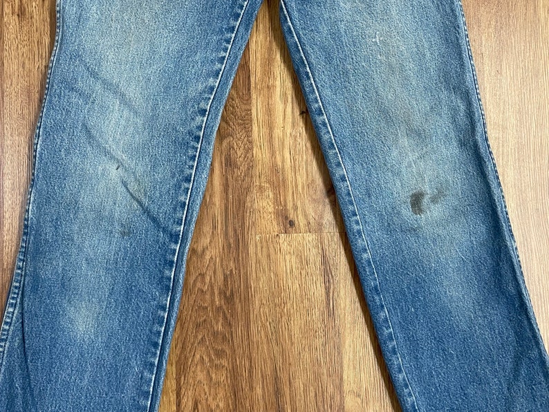 May include: A pair of blue denim jeans with visible wear and tear. The jeans have a faded appearance and are laid flat on a wooden surface.