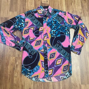 May include: A long-sleeved button-up shirt with a colorful geometric pattern. The shirt features a pink, blue, purple, and black design with a southwestern-inspired motif. The shirt has a button-down collar and a single chest pocket.