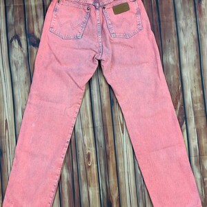 May include: Pink acid wash Wrangler jeans. The jeans have a classic five-pocket design, a brown leather patch, and the Wrangler logo on the back pockets. The jeans are laid flat on a wooden surface.