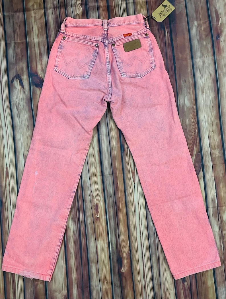 May include: Pink acid wash Wrangler jeans. The jeans have a classic five-pocket design, a brown leather patch, and the Wrangler logo on the back pockets. The jeans are laid flat on a wooden surface.