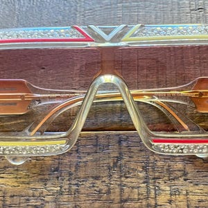 Può includere: Occhiali da sole con montatura trasparente e lenti sfumate marroni. La parte superiore della montatura presenta un design rosso, giallo e blu con strass incastonati. Le aste e il ponte sono dorati e le lenti riportano il nome del marchio.