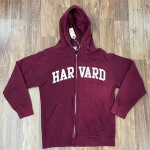 Puede incluir: Una sudadera con capucha con cremallera de color granate con la palabra "HARVARD" en letras blancas en el pecho. La sudadera tiene una capucha con cordón, una cremallera de largo completo y dos bolsillos delanteros. Una etiqueta está sujeta a la cremallera.