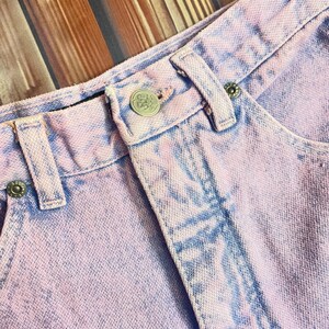 Pu&ograve; includere: Primo piano di un paio di pantaloncini di jeans lilla chiaro. I pantaloncini hanno un effetto tie-dye sbiadito con accenti blu. Il bottone e i rivetti sono argentati. Il tessuto &egrave; strutturato e mostra segni di usura.