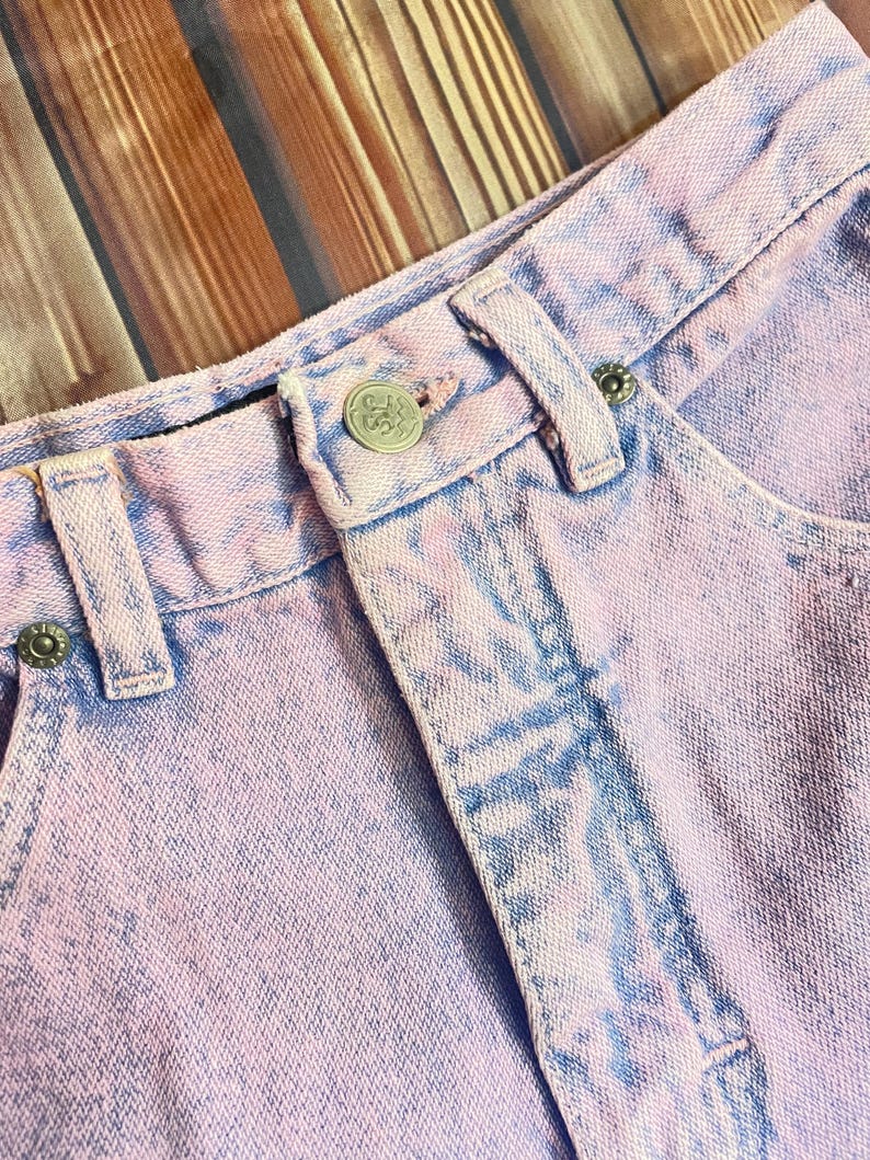Pu&ograve; includere: Primo piano di un paio di pantaloncini di jeans lilla chiaro. I pantaloncini hanno un effetto tie-dye sbiadito con accenti blu. Il bottone e i rivetti sono argentati. Il tessuto &egrave; strutturato e mostra segni di usura.