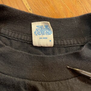 Puede incluir: Una etiqueta de cuidado blanca con texto azul unida a una camiseta negra. La etiqueta dice "Lavar a m&aacute;quina, secar en secadora, detergente suave, temperatura baja, sin lej&iacute;a con cloro, no planchar el emblema, no limpiar en seco, RN 20100, H".
