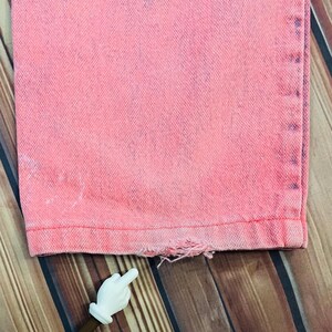 May include: Close-up of the bottom hem of pink denim jeans. The jeans have a distressed, faded look with a frayed hemline. The fabric appears to be a medium weight denim. The jeans are displayed on a wooden surface.