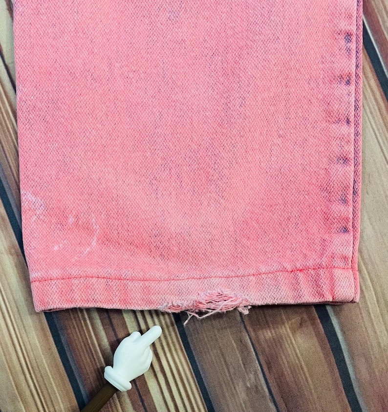 May include: Close-up of the bottom hem of pink denim jeans. The jeans have a distressed, faded look with a frayed hemline. The fabric appears to be a medium weight denim. The jeans are displayed on a wooden surface.