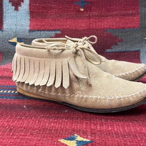 Könnte beinhalten: Ein Paar beige Wildleder-Mokassin-Stiefel mit Fransen. Die Stiefel haben ein Schnürdesign und eine genähte Sohle. Das Schuhwerk wird auf einem gemusterten rot-blauen Teppich präsentiert.